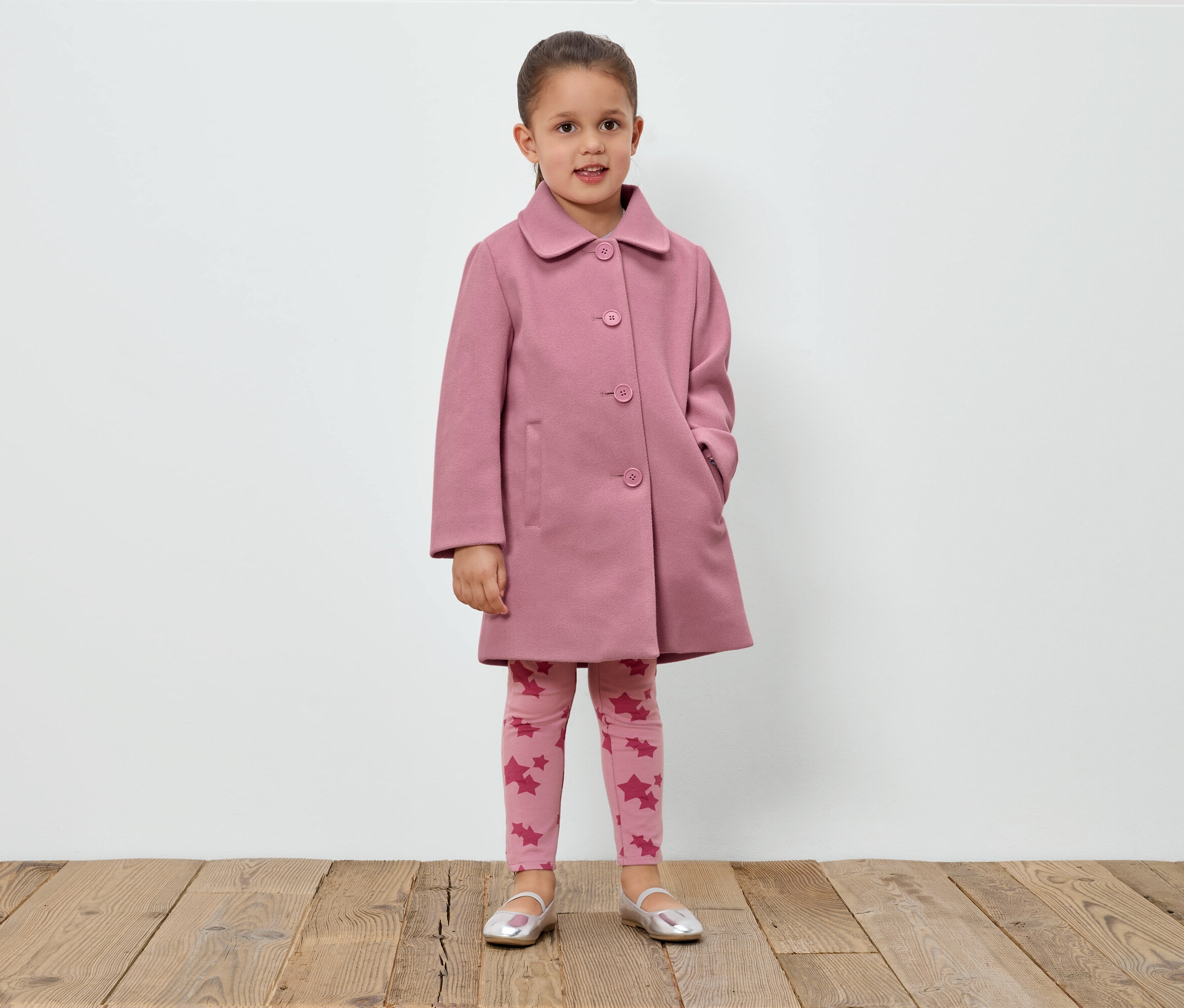 Dívka stojí před bílou zdí a má na sobě růžový kabát, legíny s hvězdami a stříbrné boty.