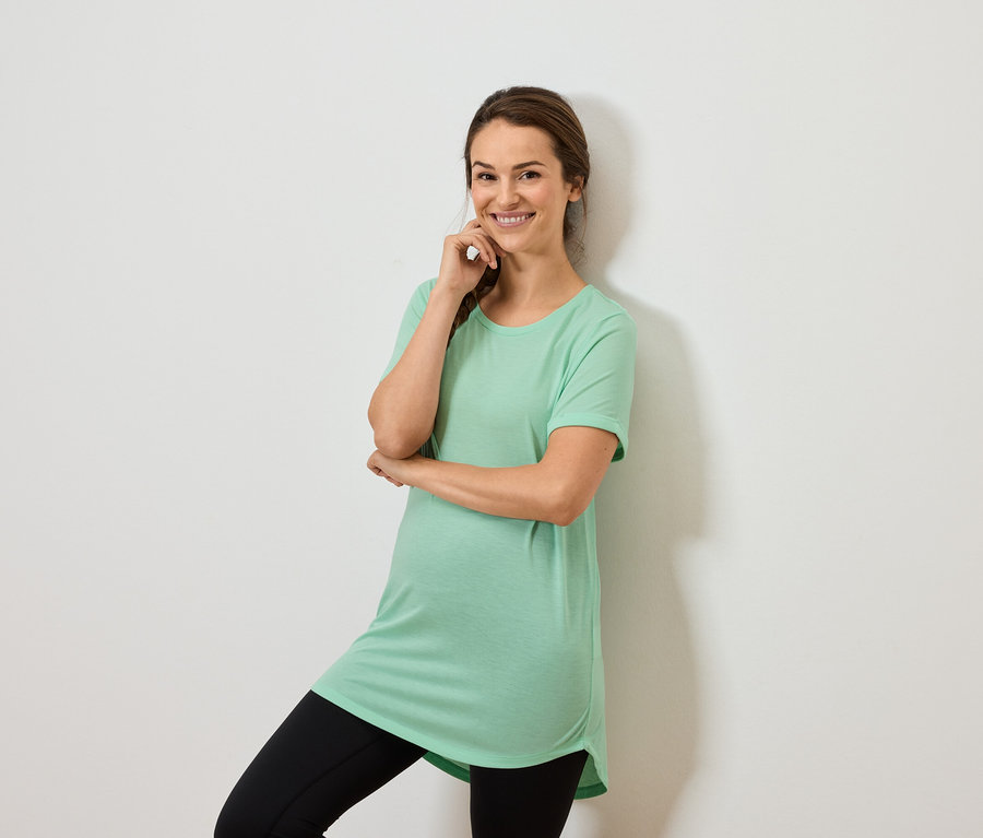 Žena v mátově zeleném topu a černých legínách pózuje s rukou na bradě.