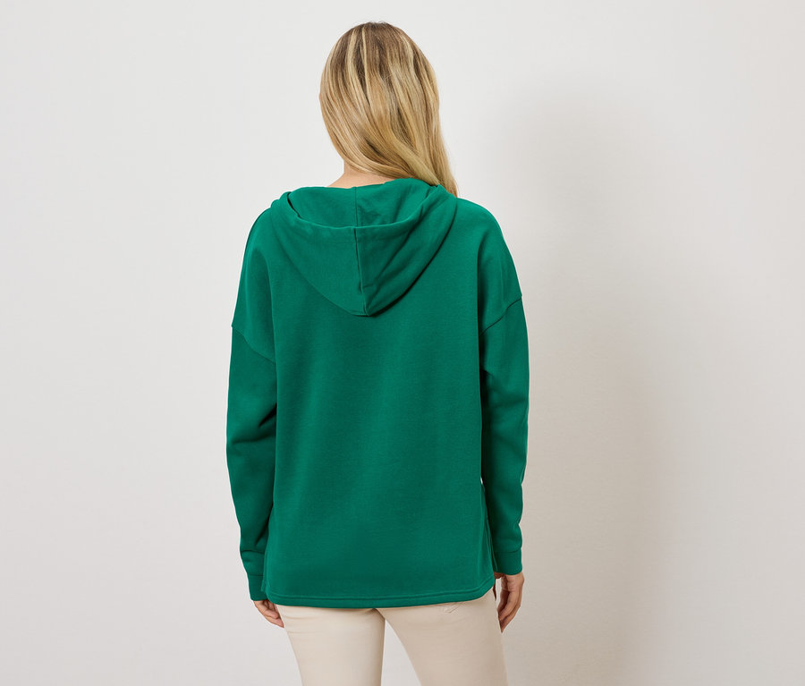 Žena zády k fotoaparátu má na sobě zelenou mikinu s kapucí a světlé kalhoty.