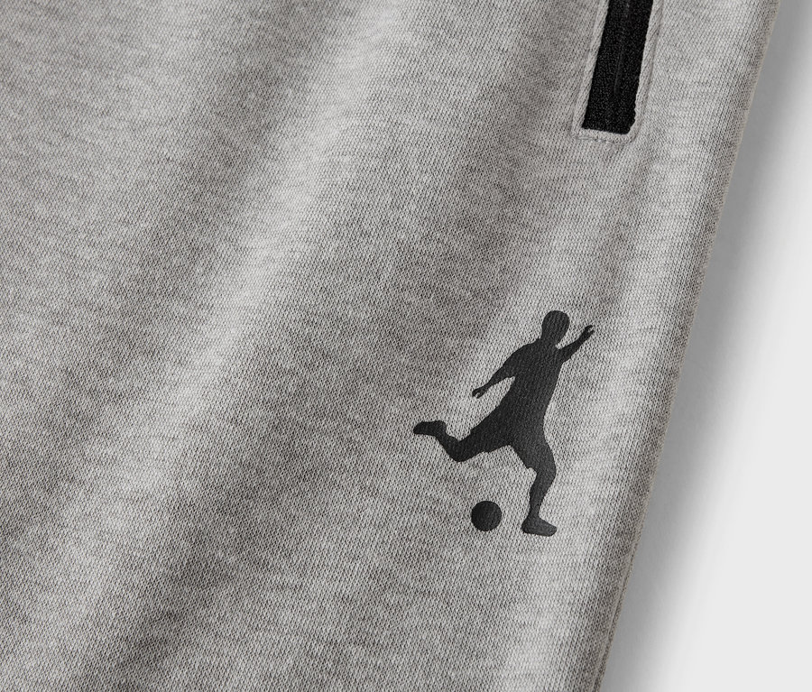 Detail šedých tepláků s černou siluetou fotbalisty a kapsou na zip.