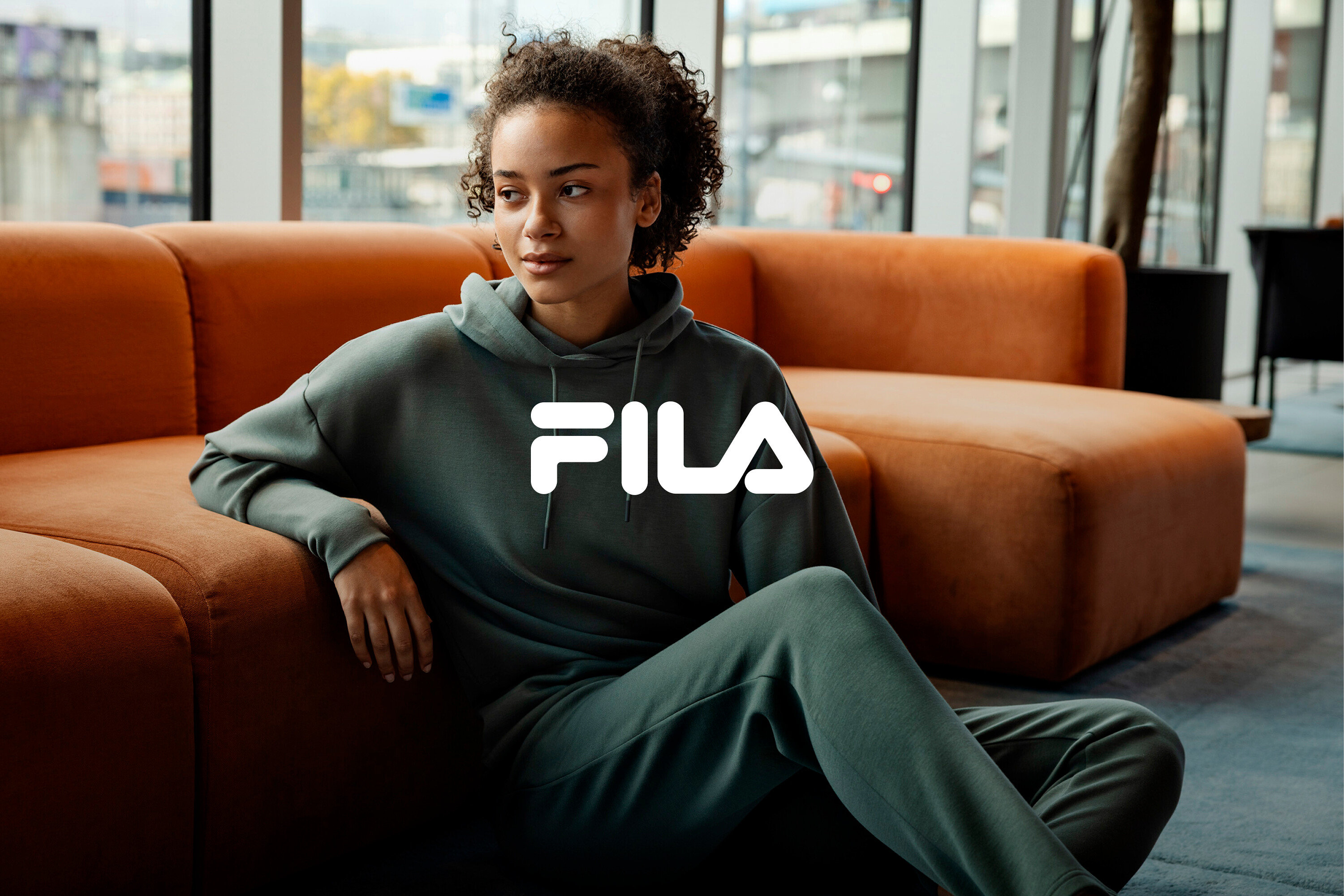 Žena sedí na zemi, opřená o oranžovou pohovku, a má na sobě zelenou teplákovou soupravu Fila.