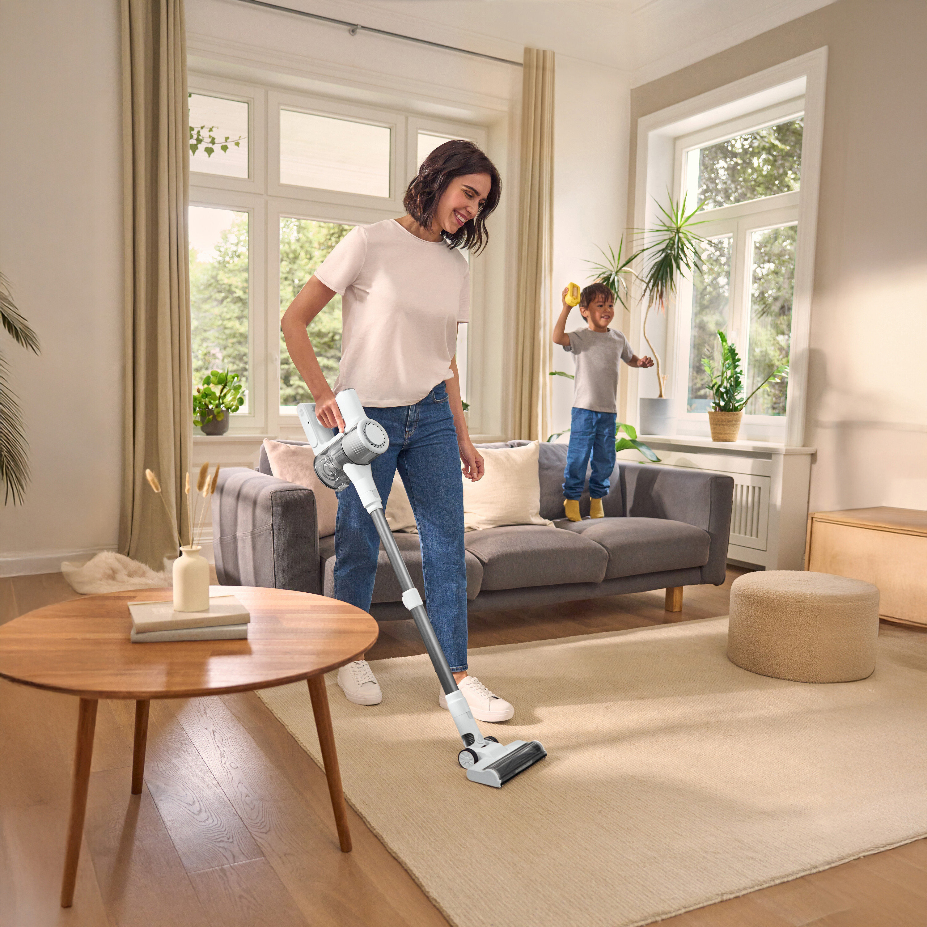 Žena vysává koberec akumulátorovým vysavačem, dítě stojí na pohovce. Kulatý stůl vlevo, okno v pozadí.