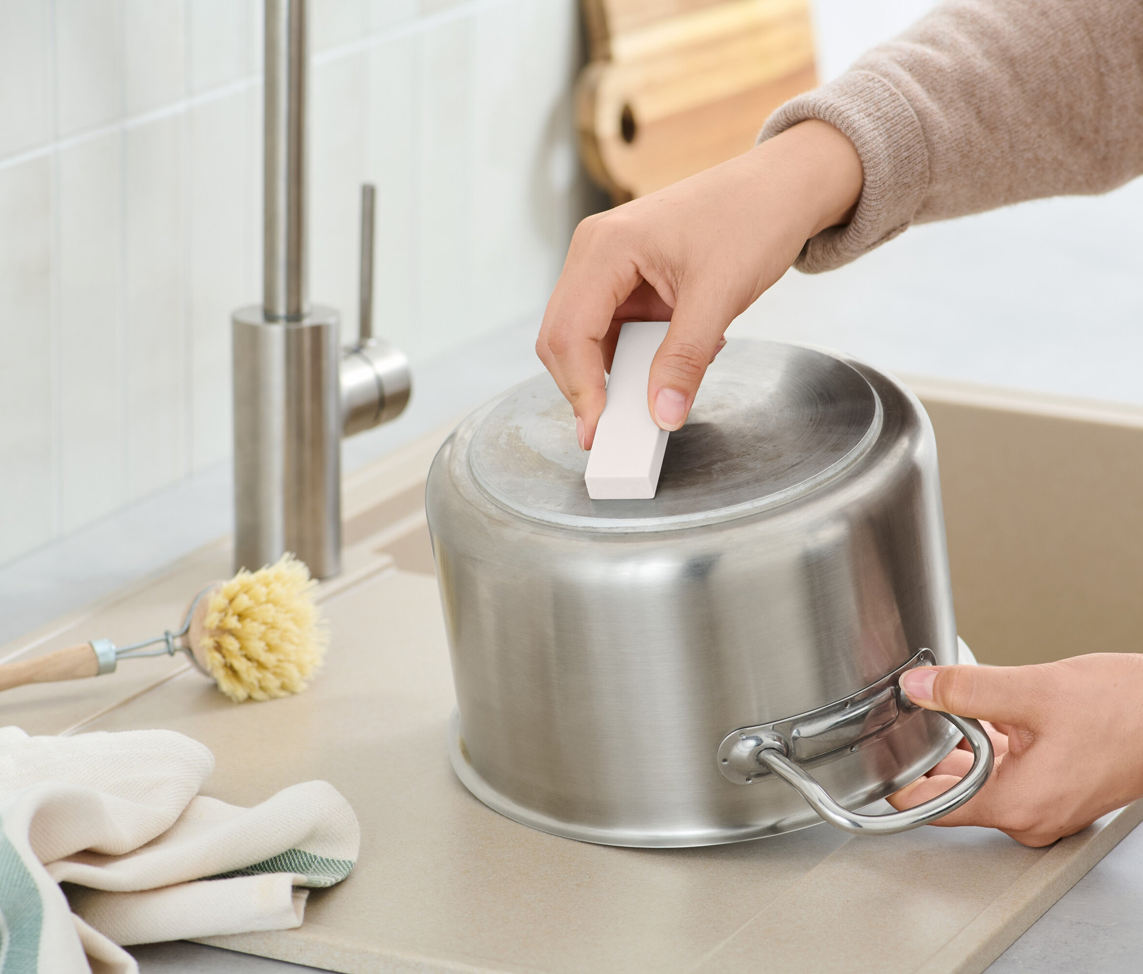 Osoba čistí nerezový hrnec čisticí houbičkou v umyvadle, vedle kartáče a hadříku.