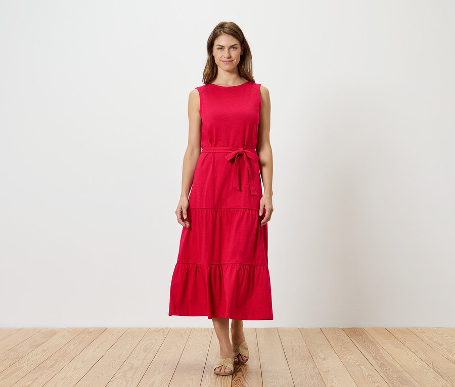 Žena stojí před bílou stěnou a má na sobě červené šaty bez rukávů s volány a páskem a sandály.