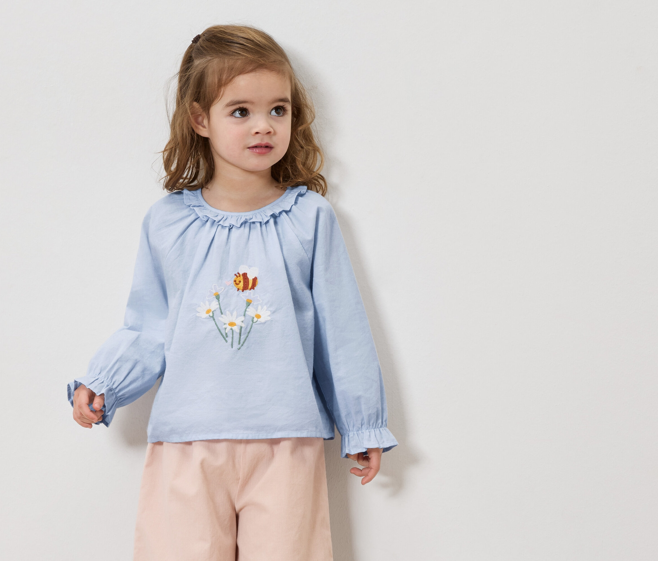Dívka s blond vlasy má na sobě světle modrou halenku s výšivkou sedmikrásek a včel a růžové kalhoty.
