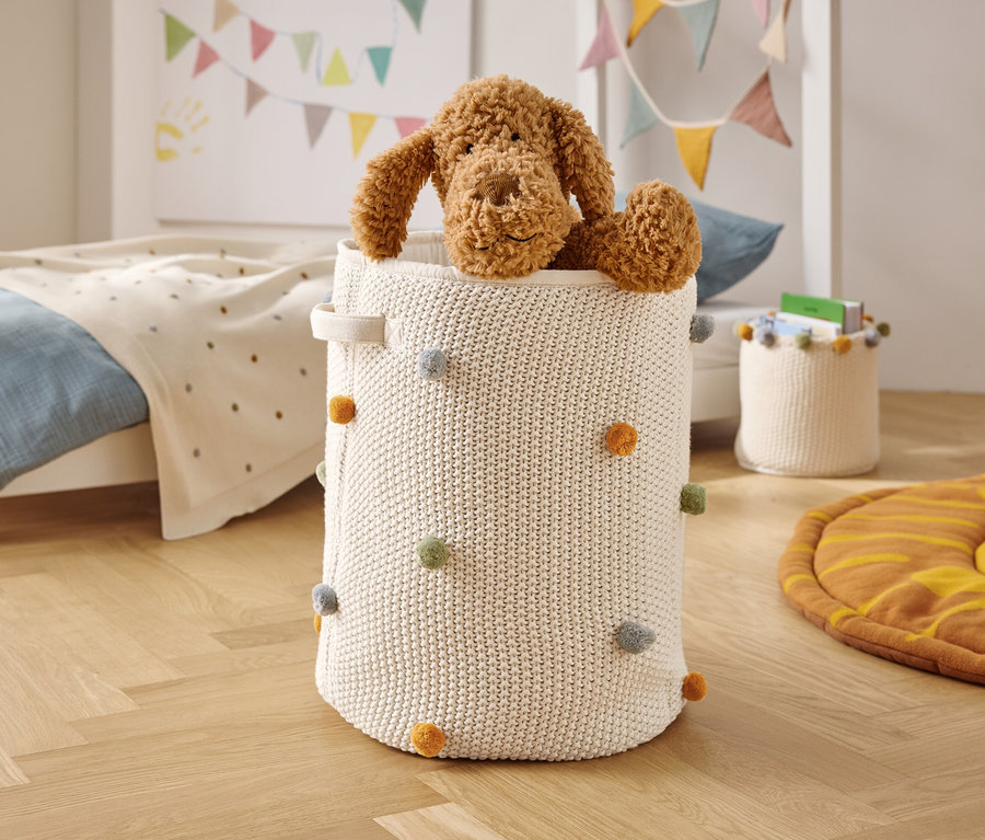 Hnědý plyšový pes sedí v bílém pleteném koši s barevnými bambulkami.