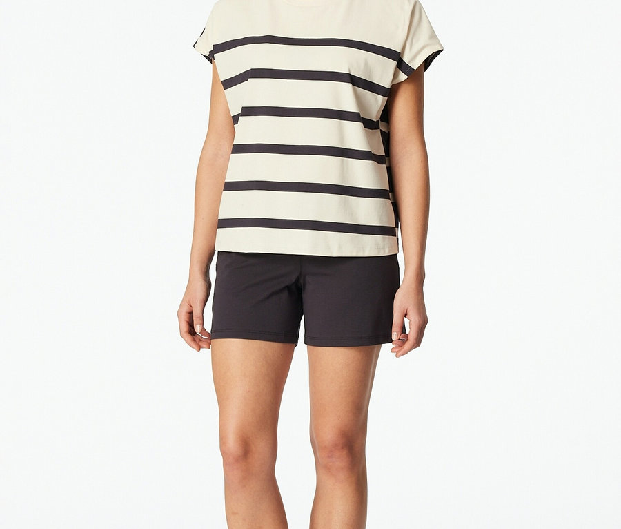 Žena stojí bosá na bílém pozadí v pruhovaném topu a černých šortkách.