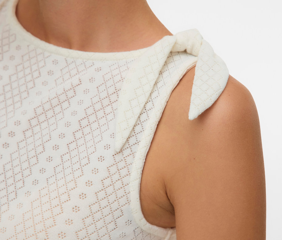 Detail ženy v bílém děrovaném tílku s mašlí na rameni.