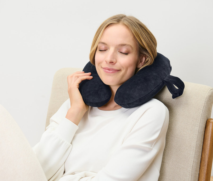 Žena sedí se zavřenýma očima na židli a má na sobě tmavě modrý cestovní polštář.