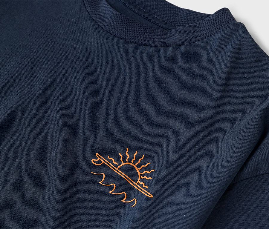 Detailní záběr na tmavě modré tričko s oranžovým potiskem slunce nad surfovým prknem a vlnami.