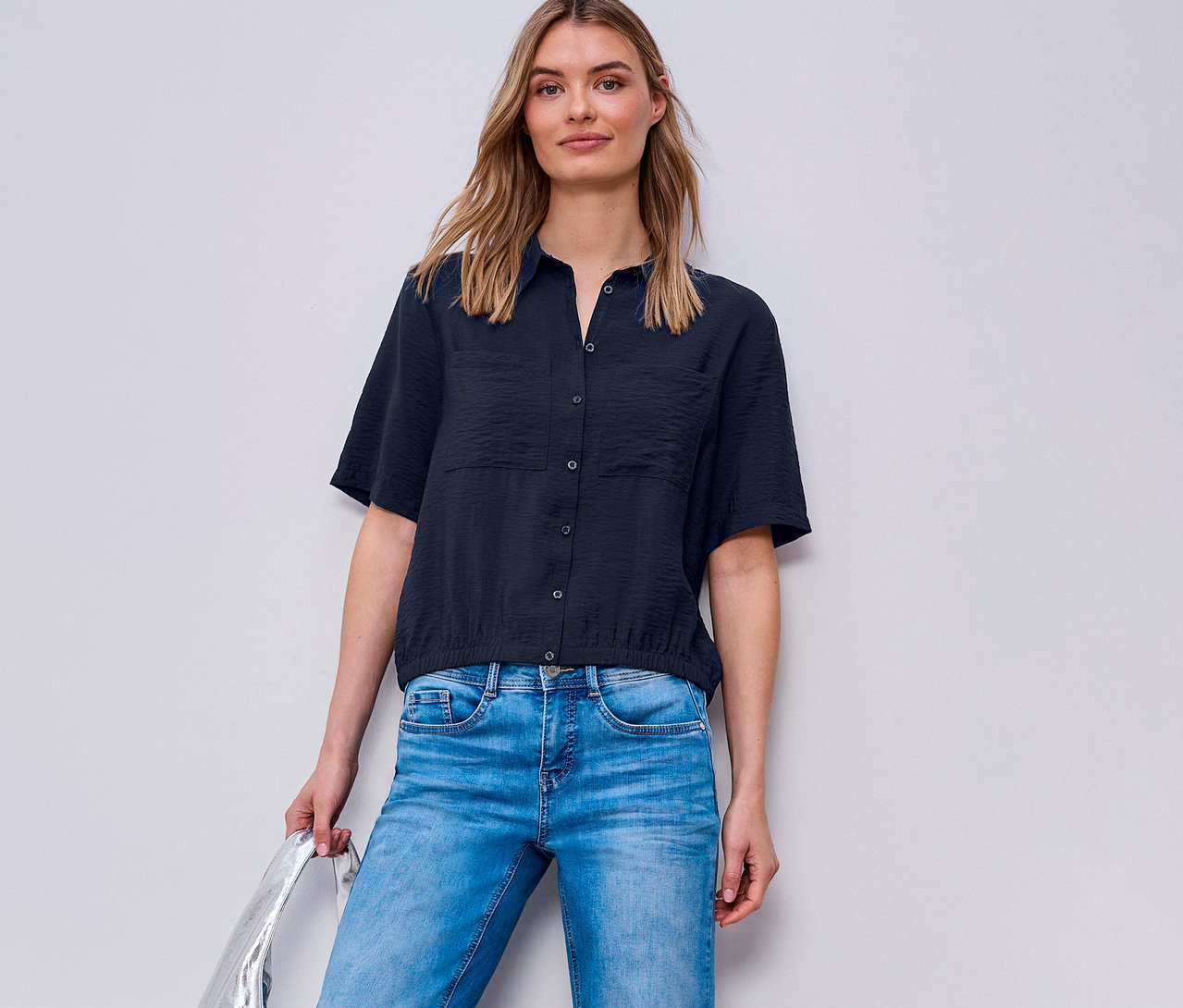 Žena má na sobě džíny a košilovou halenku Street One. Drží stříbrnou tašku.