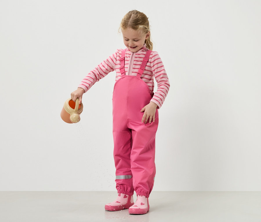 Dívka lije vodu z konve, má na sobě růžové lacláče a boty.