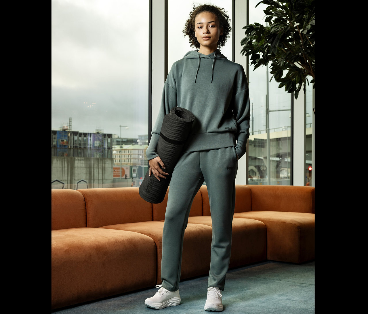 Mladá žena v šedém sportovním oblečení drží srolovanou podložku na jógu před oknem.