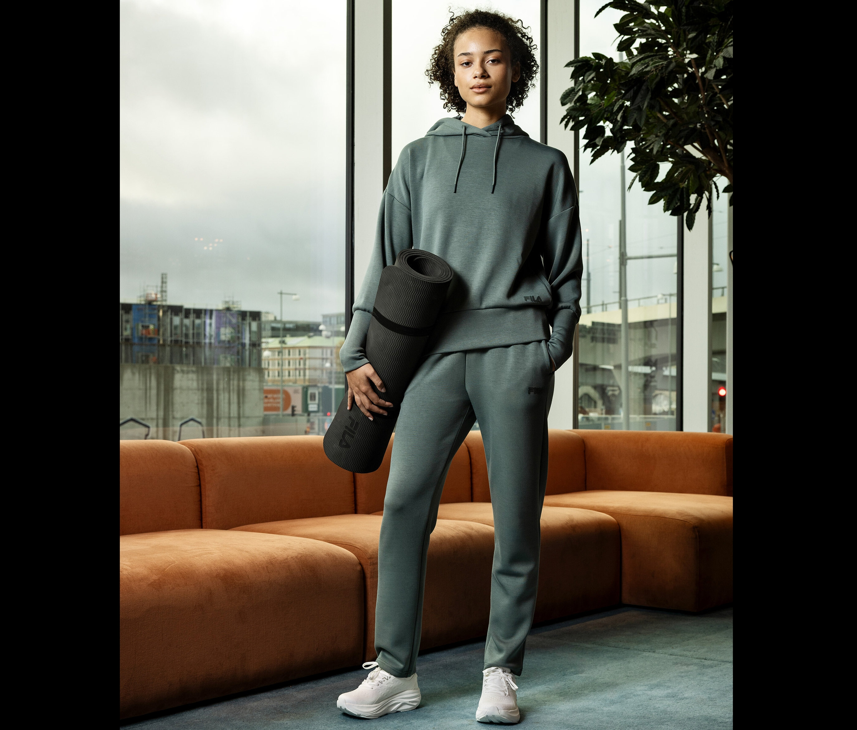 Mladá žena v šedém sportovním oblečení drží srolovanou podložku na jógu před oknem.