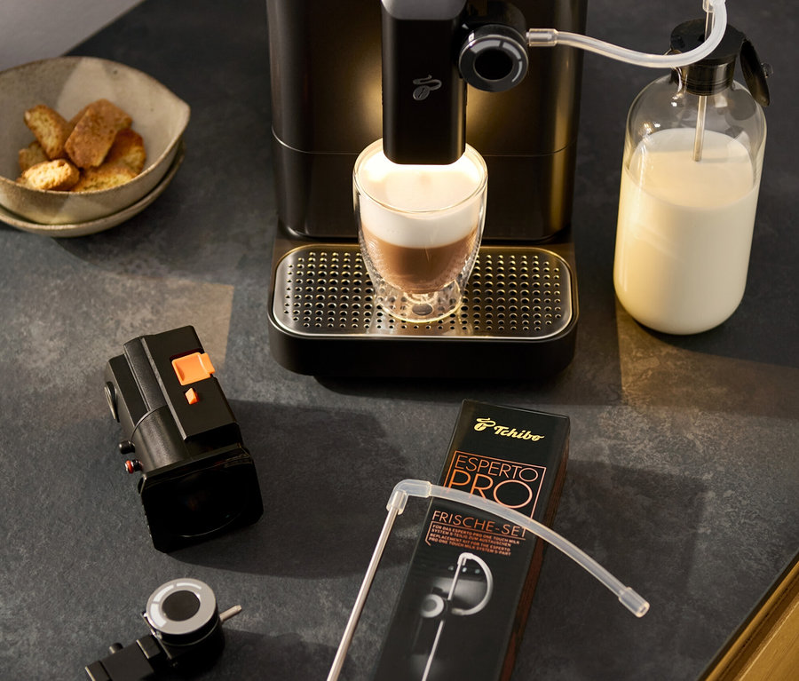 Kávovar »Esperto 2 Milk« Granite Black připravuje latte macchiato. Vedle stojí otočný regulátor mléčné pěny, miska s pečivem a balení sady příslušenství.