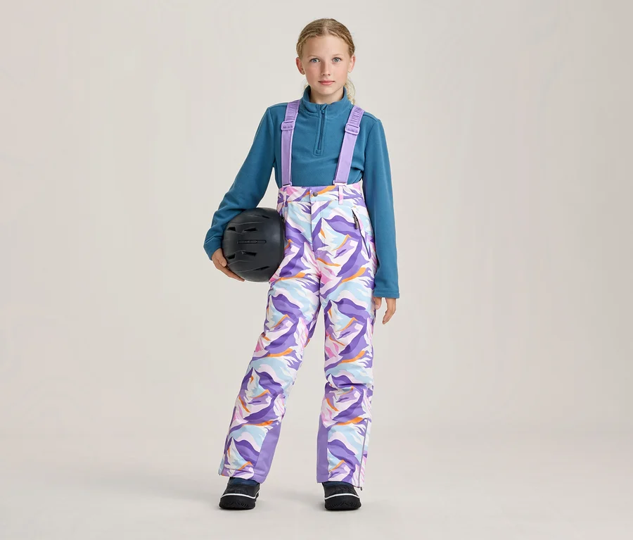 Mladá dívka v tyrkysovém fleecovém topu a barevných vzorovaných lyžařských kalhotách s fialovými šlemi, držící černou lyžařskou helmu v levé ruce.