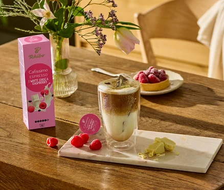 Na dřevěném stole stojí sklenice s kávou, vedle ní leží kousky bílé čokolády a maliny na mramorovém prkénku. V pozadí je vidět balení Cafissimo Flavoured Espresso – White Choc & Raspberry – 10 kapslí.