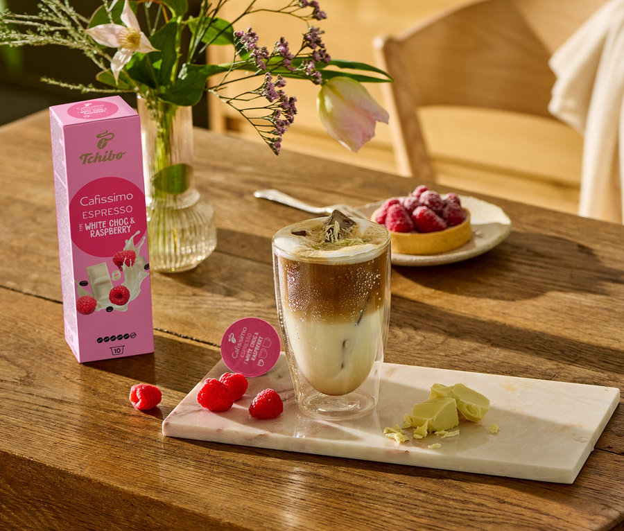 Na dřevěném stole stojí sklenice s kávou, vedle ní leží kousky bílé čokolády a maliny na mramorovém prkénku. V pozadí je vidět balení Cafissimo Flavoured Espresso – White Choc & Raspberry – 10 kapslí.