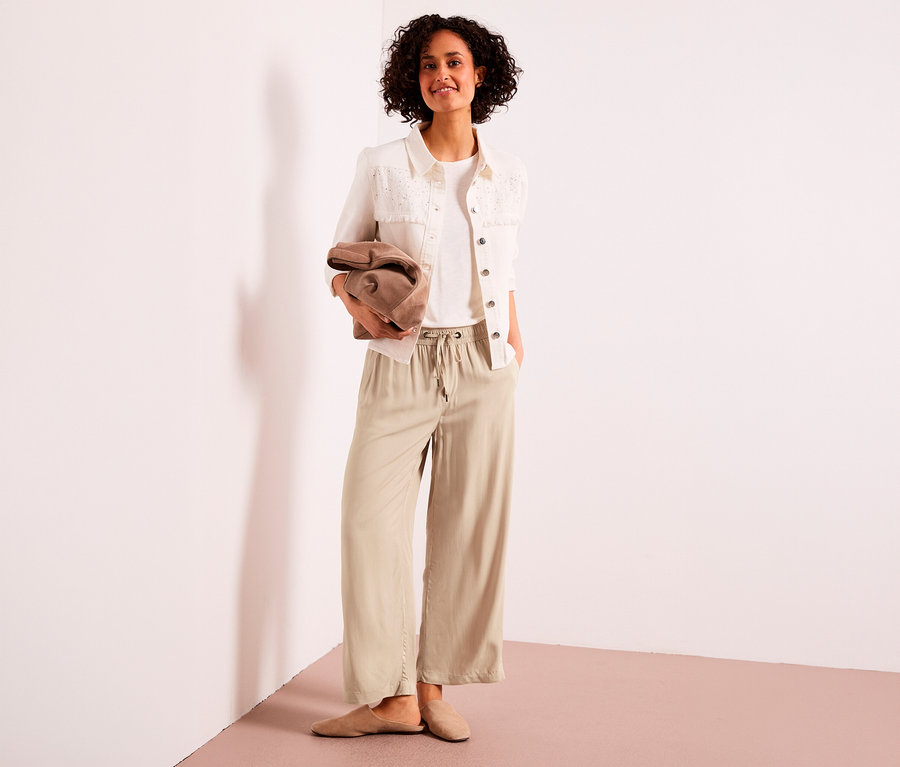 Usmívající se žena stojí, má na sobě bílé tričko, bílou bundu a béžové 7/8 kalhoty culottes Street One, drží hnědou kabelku.