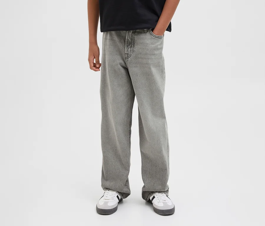 Osoba v šedých džínách Jack & Jones JJOriginal Alex a bílých teniskách. Jedna ruka v kapse.