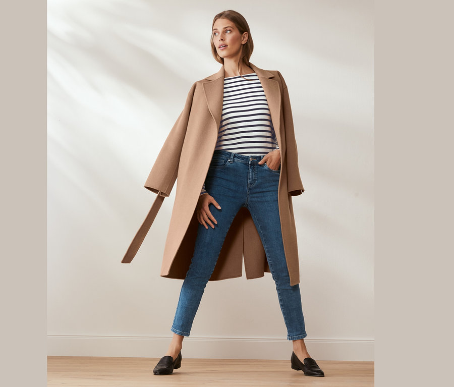 Žena pózuje ve vlněném kabátě s kašmírem, modrých džínách a pruhovaném tričku.
