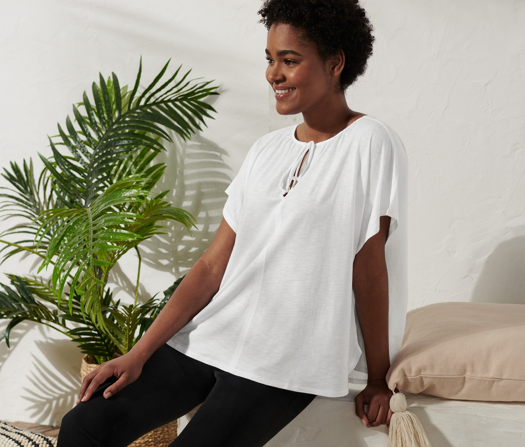 Žena sedí a usmívá se, má na sobě halenkové triko se šňůrkou na zavazování, černé kalhoty a sandály na platformě.