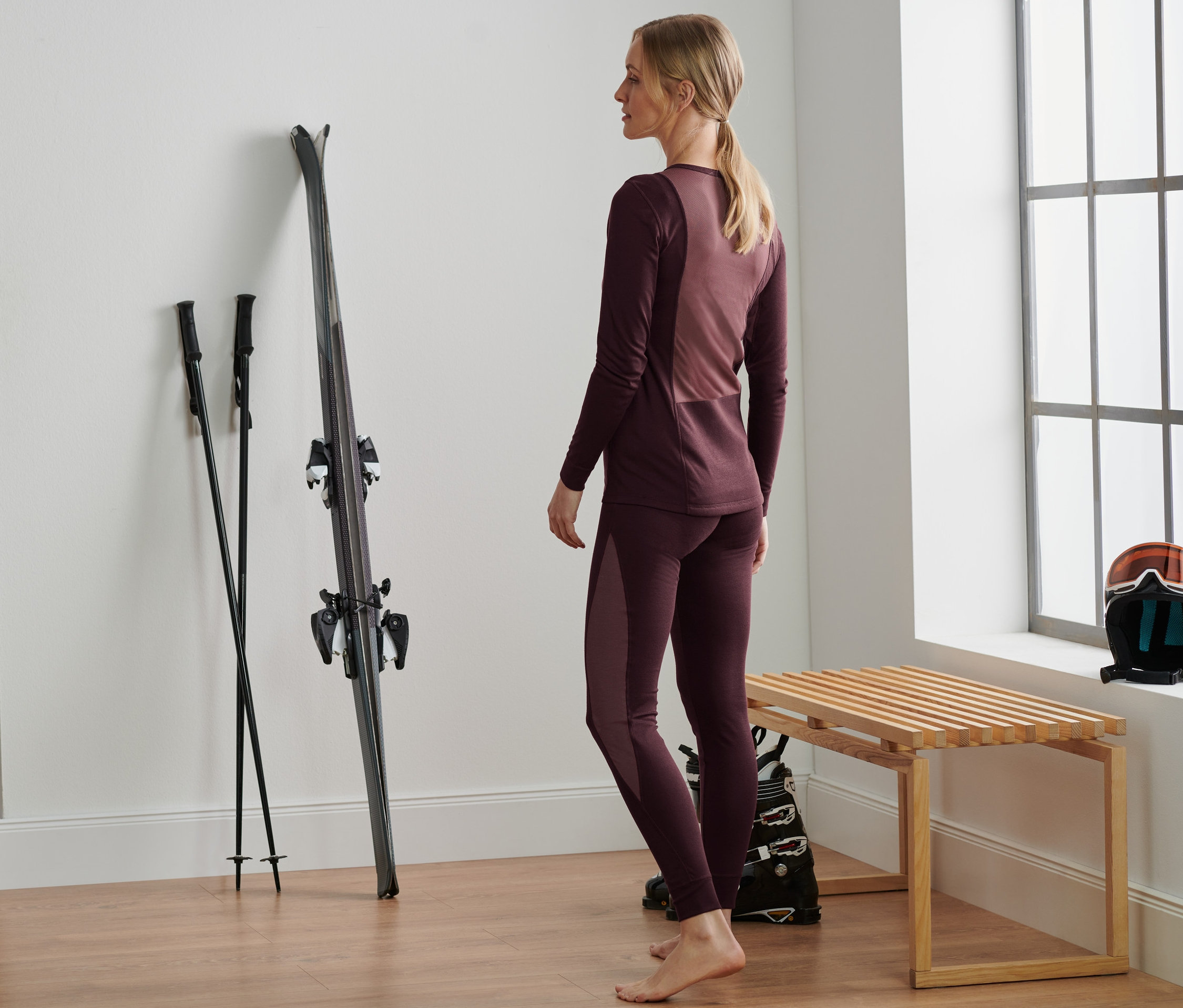 Žena stojí v bordó funkčním spodním prádle vedle lyží a hůlek.
