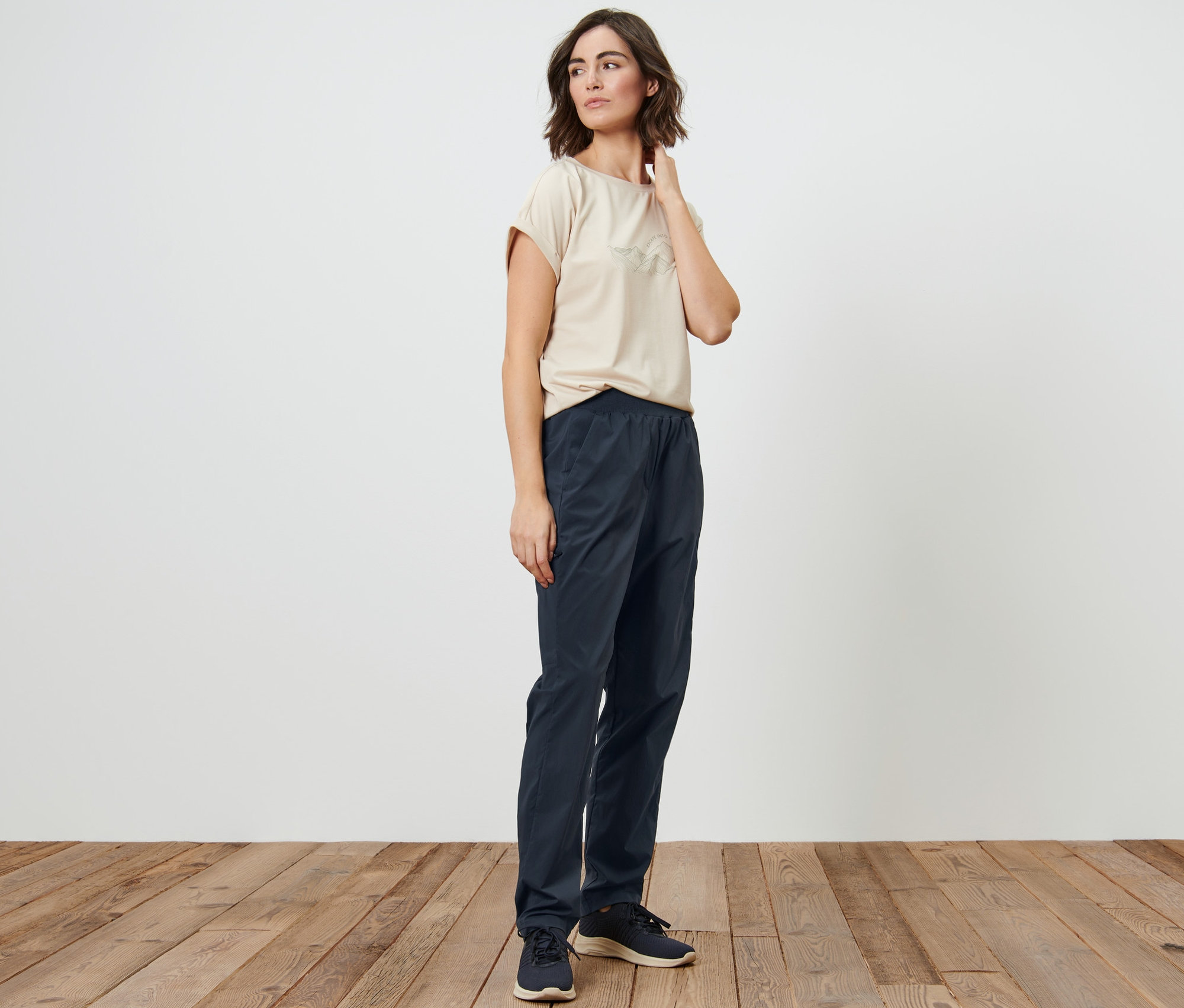 Žena pózuje v béžovém tričku a tmavě modrých funkčních turistických kalhotách.