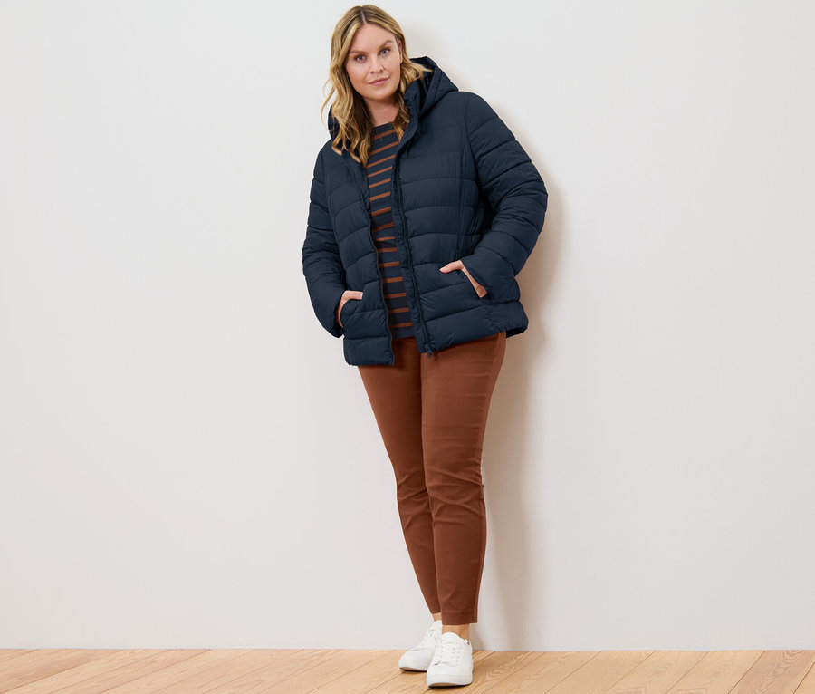 Žena pózuje v tmavě modré prošívané bundě s kapucí, proužkovaném tričku s 3/4 rukávy a koňakových strečových kalhotách.