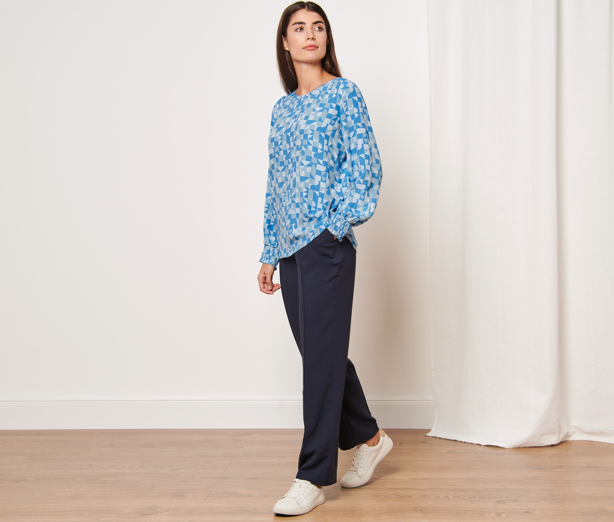 Žena pózuje v modré halence s potiskem a tmavě modrých kalhotách.