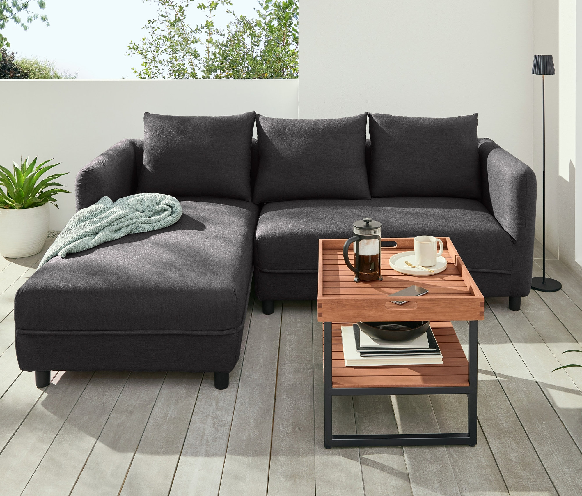 Černá pohovka stojí na terase vedle LED stojací lampy a konferenčního stolku s odnímatelným podnosem.