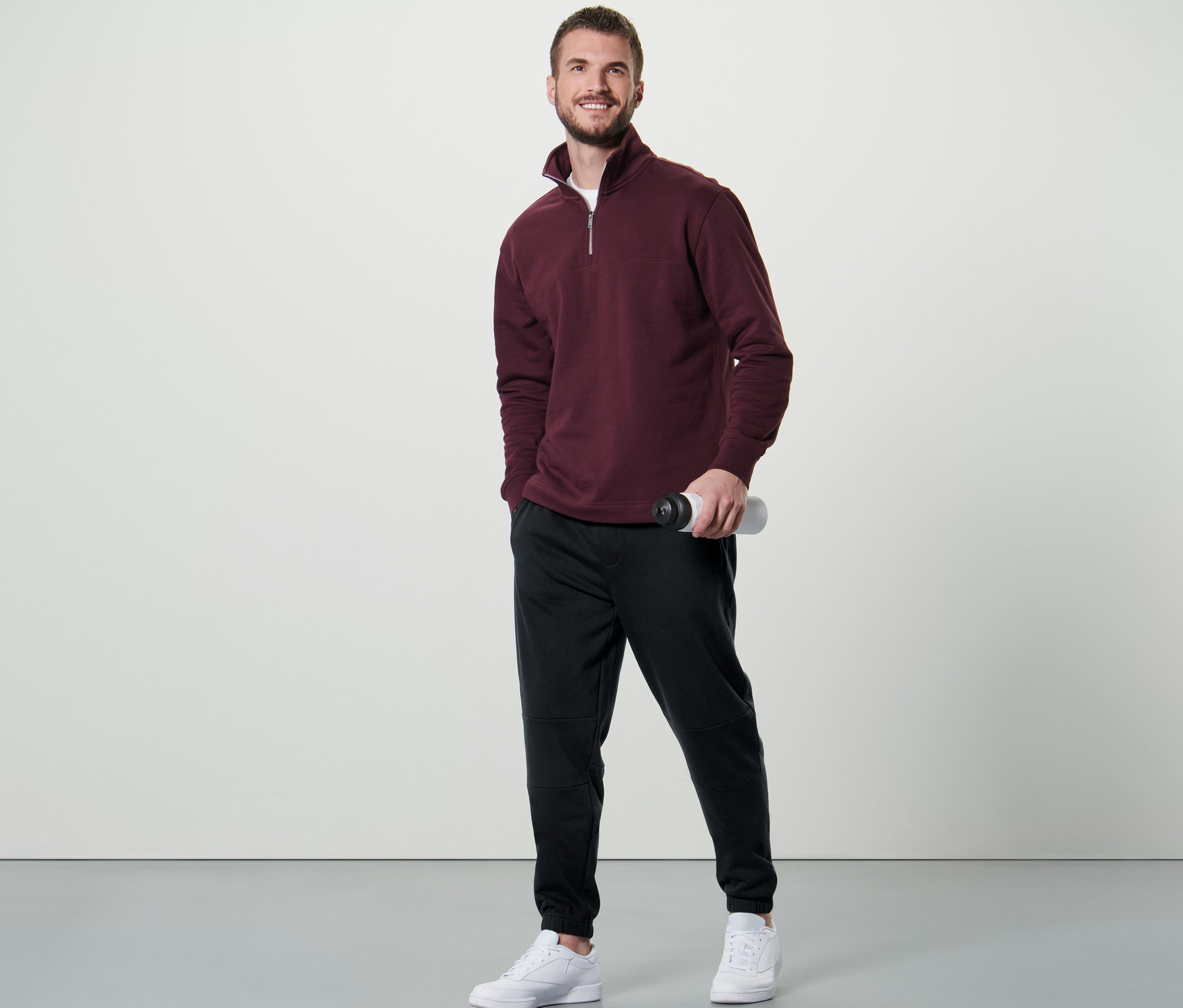 Muž stojí a drží láhev na vodu, má na sobě teplákový svetr troyer a teplákové kalhoty.