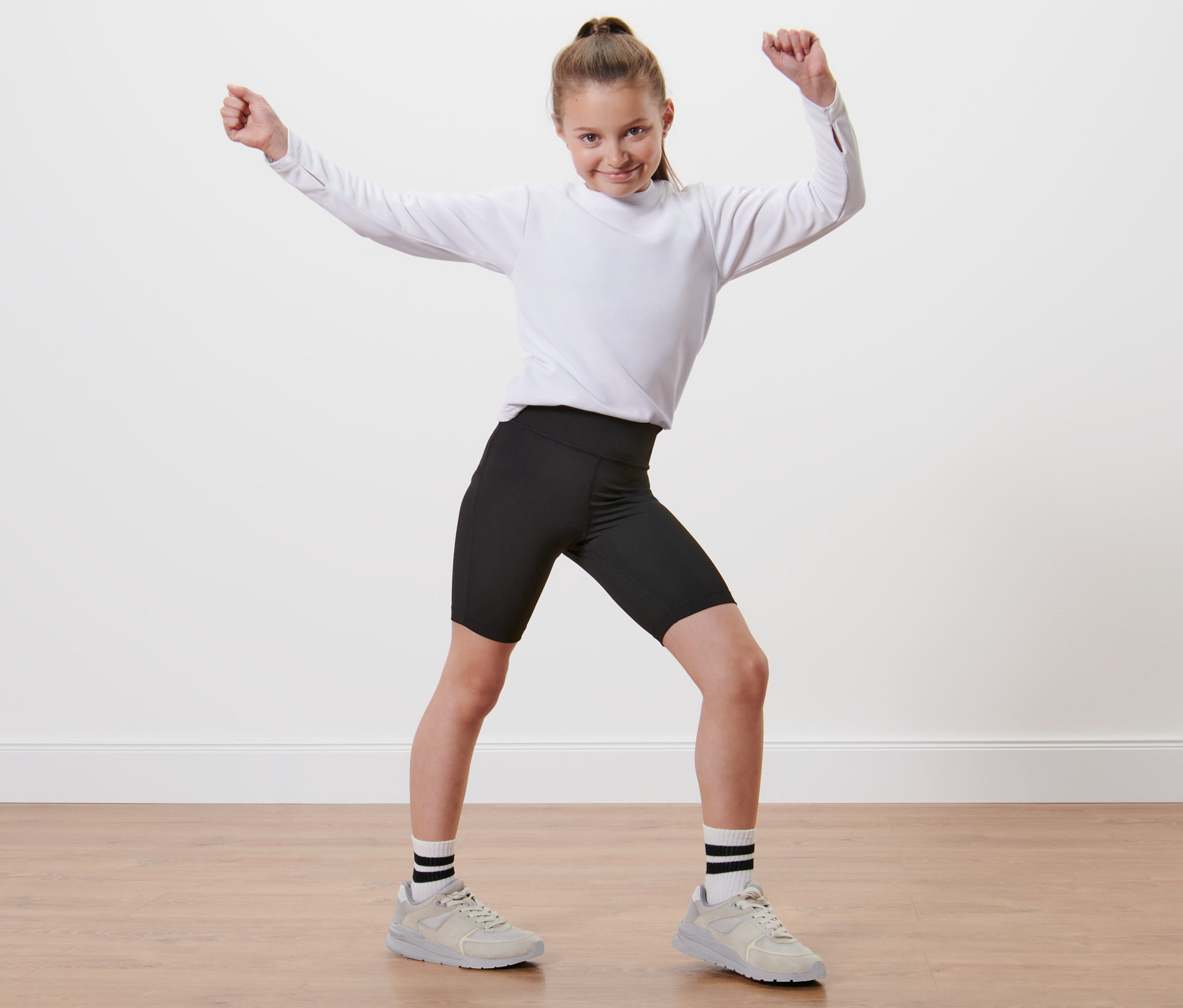 Dívka v bílém tričku a černých sportovních šortkách 2 ks stojí s rukama nahoře.
