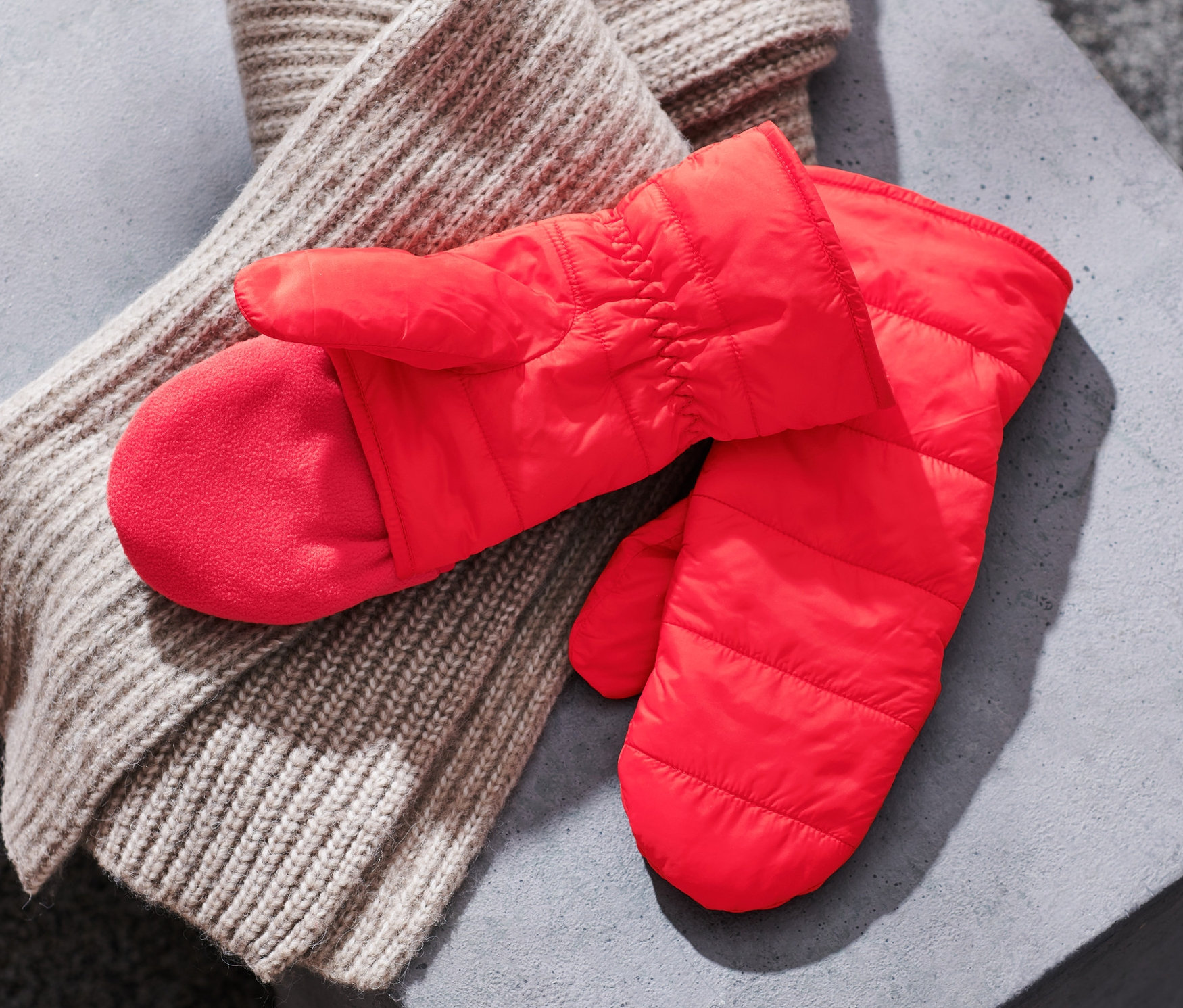 Na šedé ploše leží pár červených prošívaných odklápěcích palčáků a béžová pletená šála.