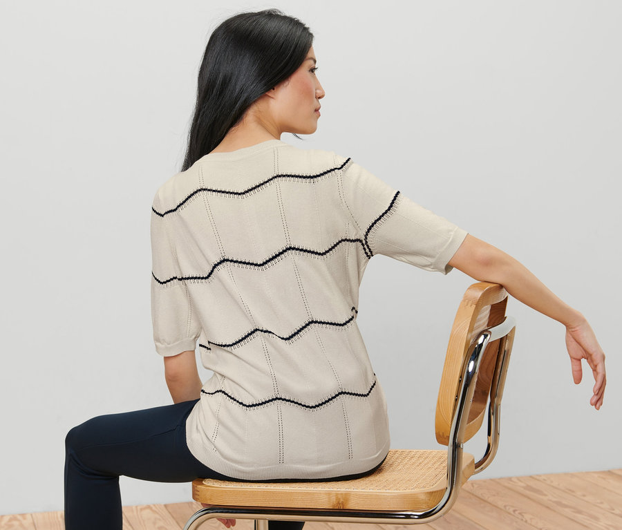 Žena sedí na židli zády ke kameře a má na sobě kardigan z jemného úpletu s ažurovým vzorem.