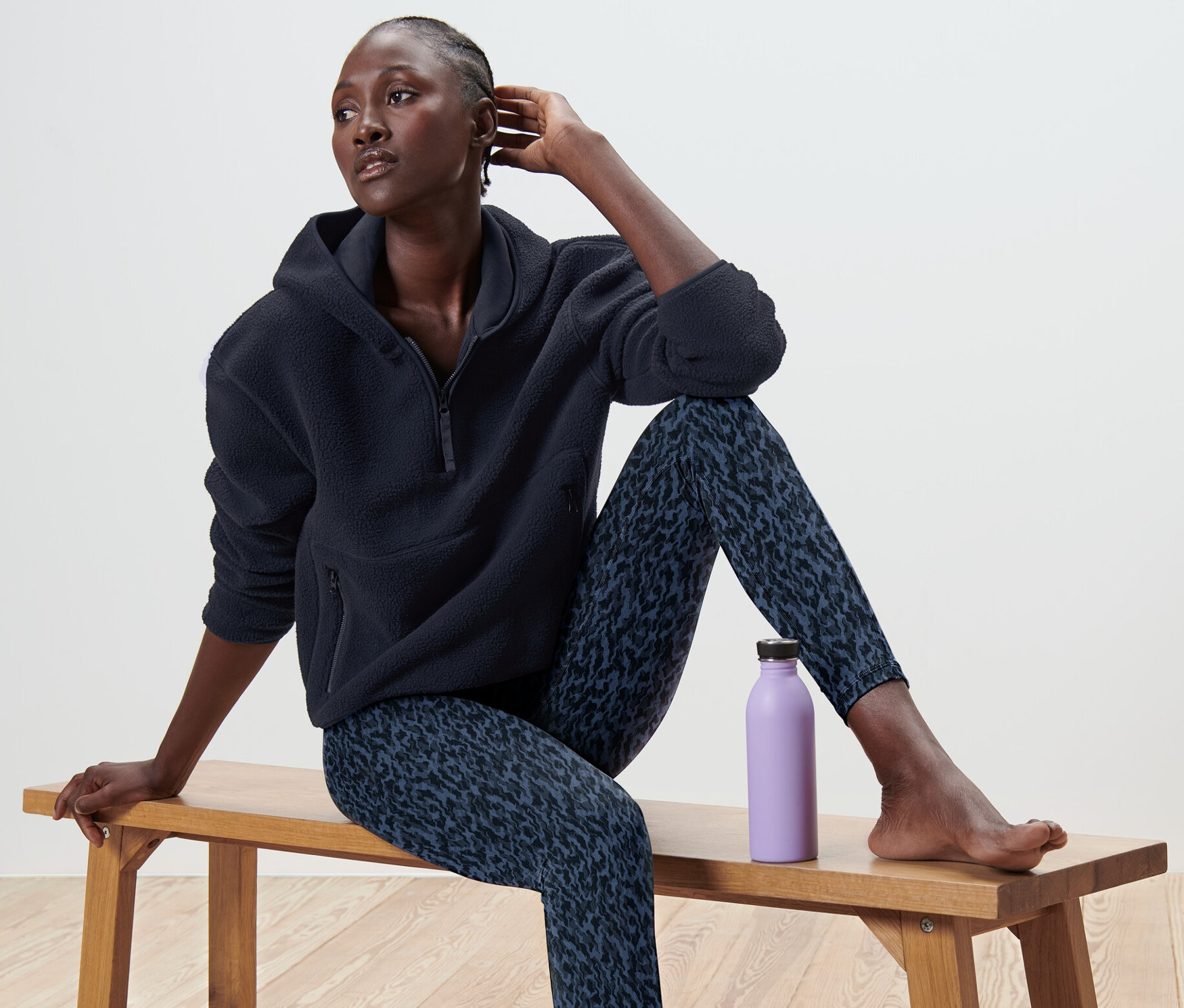 Žena sedí na lavičce v plyšovém svetru s kapucí a vzorovaných legínách, vedle ní je fialová láhev.