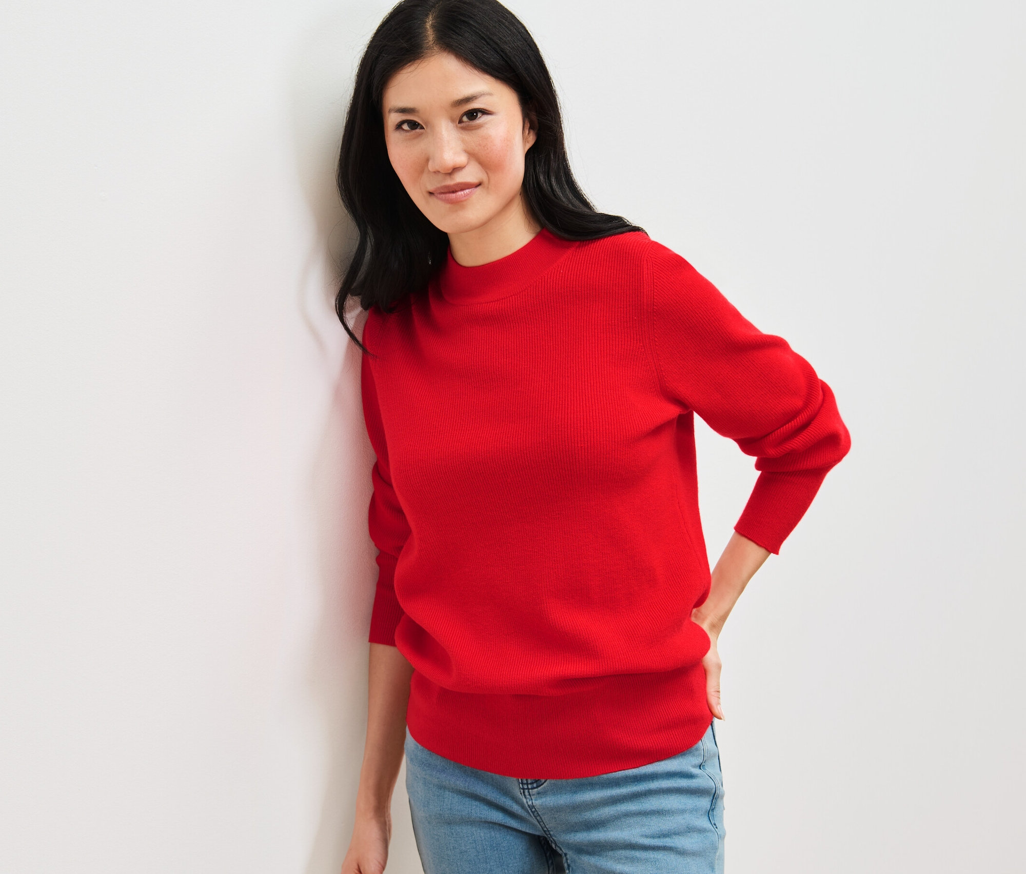 Žena opřená o zeď má na sobě červený svetr z jemného úpletu a džíny.