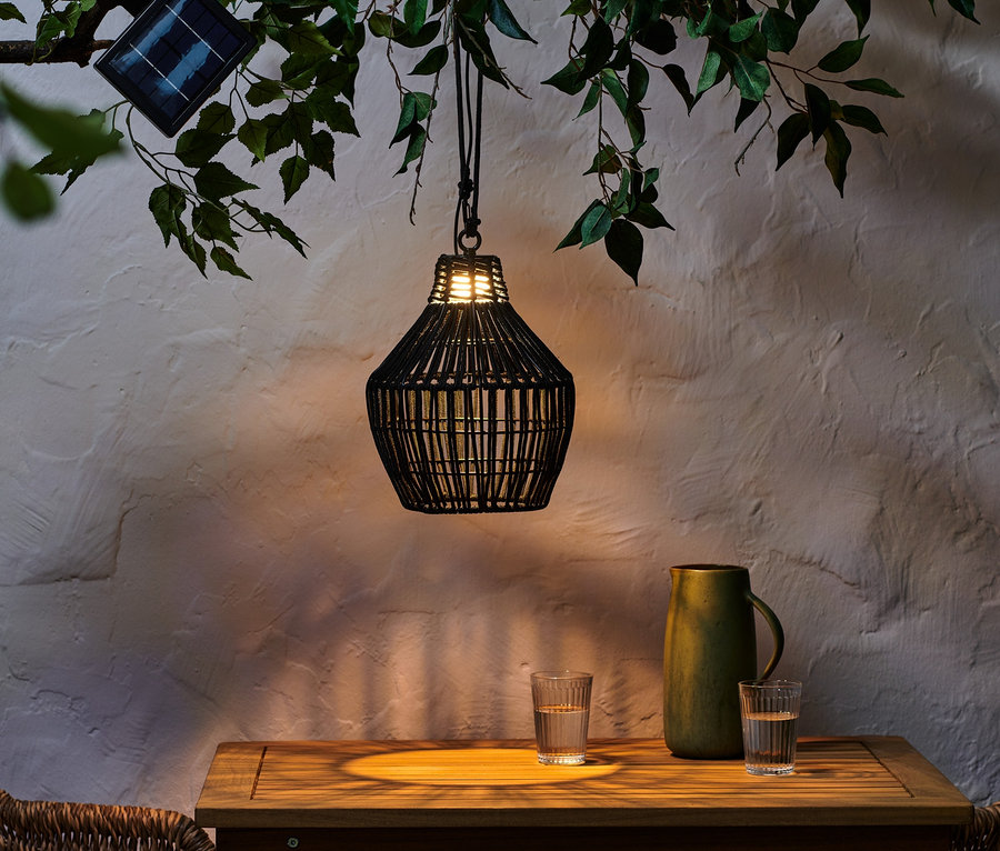 Černé solární závěsné svítidlo s textilním páskem visí z větve nad stolem se sklenicemi a zeleným džbánem.
