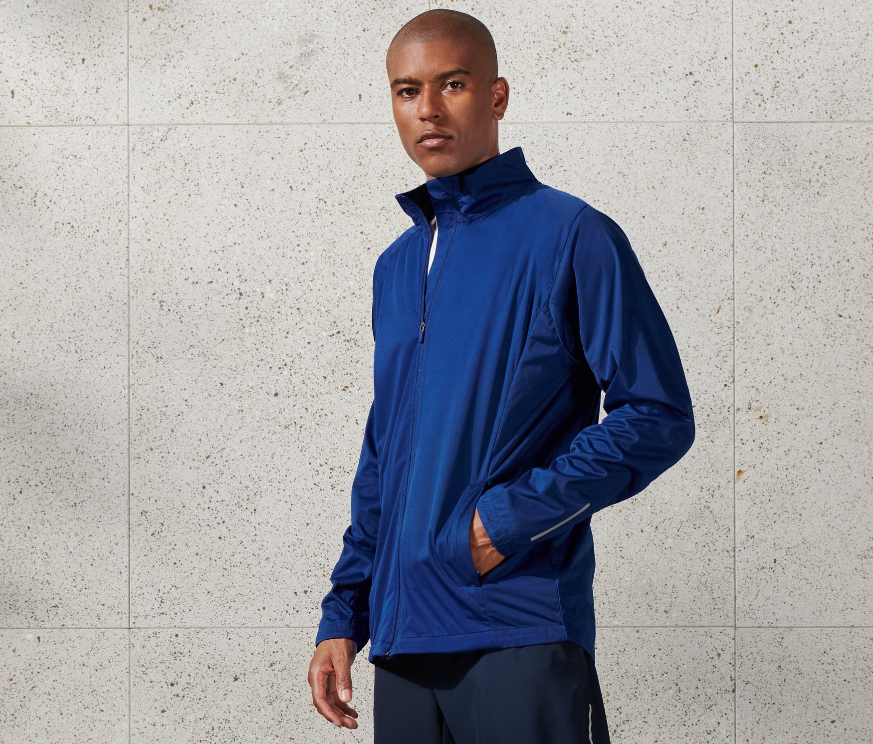 Muž má na sobě modrou větruvzdornou funkční bundu, šortky a bílé tenisky.