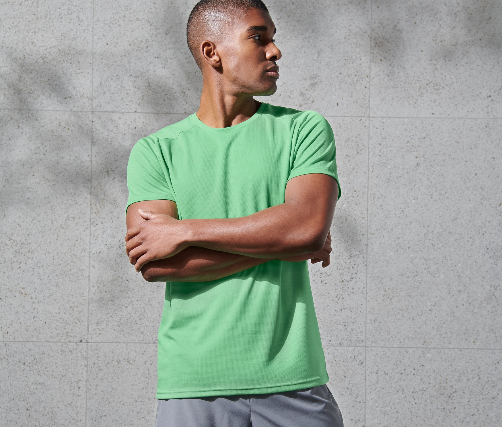 Muž se zkříženýma rukama má na sobě zelené funkční triko a šedé šortky.