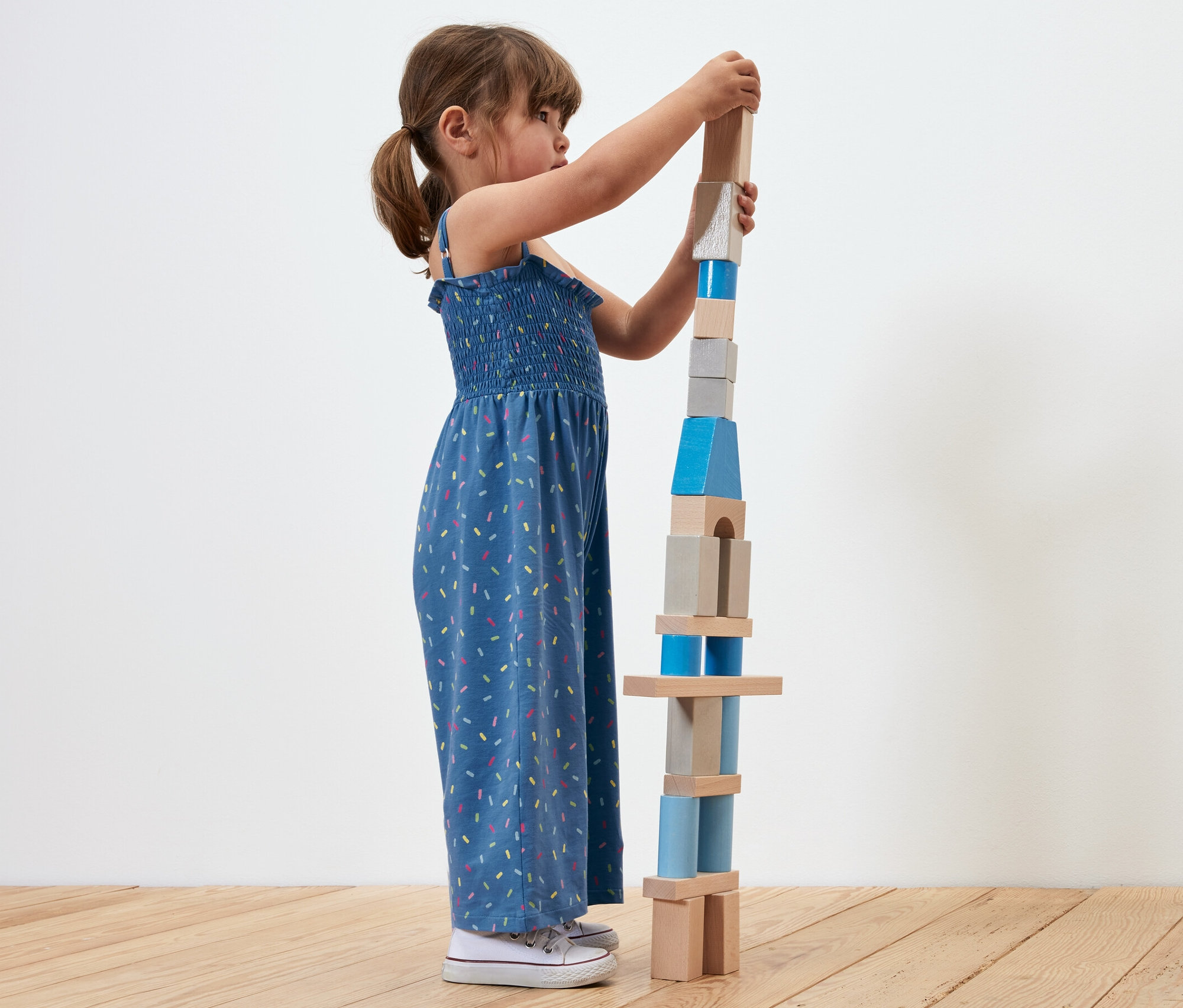 Dívka staví věž z dřevěných bloků. Má na sobě modrý overal v 7/8 délce.