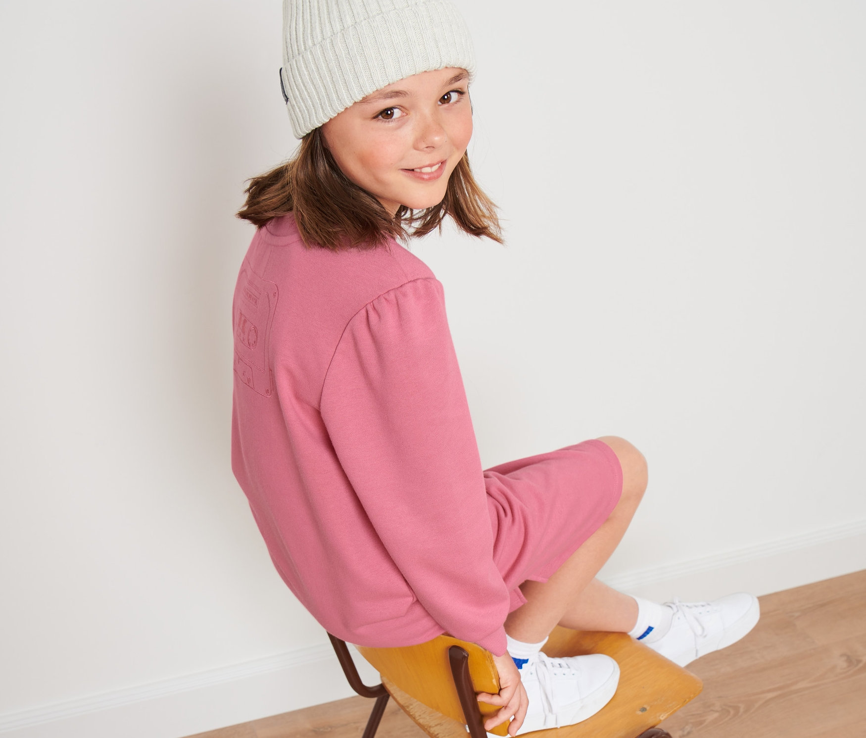 Dívka sedí na židli a má na sobě dětské teplákové šaty a bílou čepici.