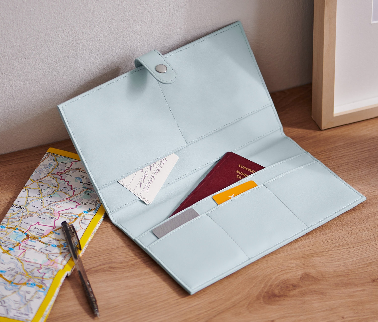 Otevřený světle modrý cestovní organizér leží na dřevěném stole vedle mapy a pera.
