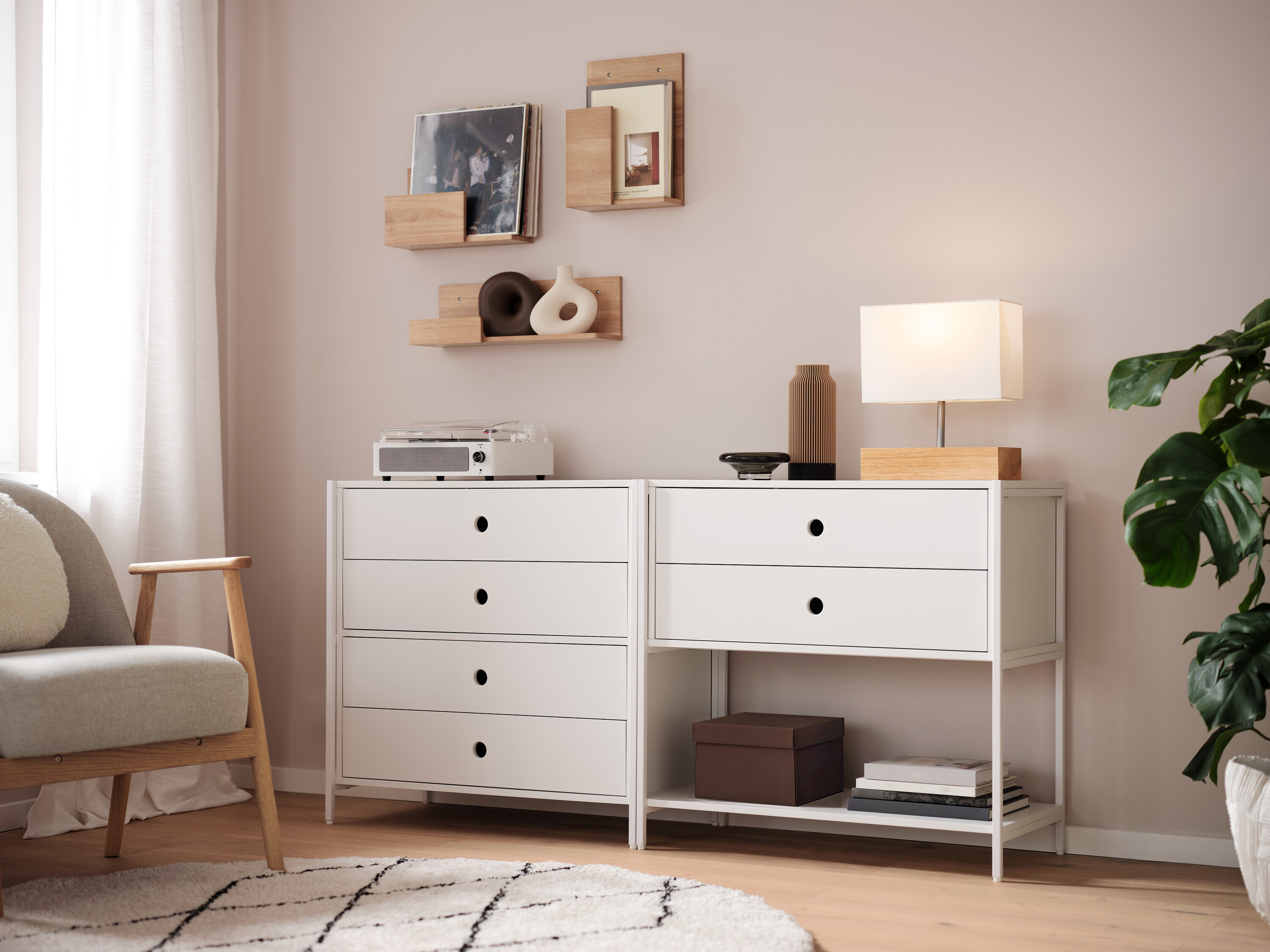 Dva bílé prádelníky stojí vedle sebe, na nich je lampa, váza a rádio. Na zdi jsou police.