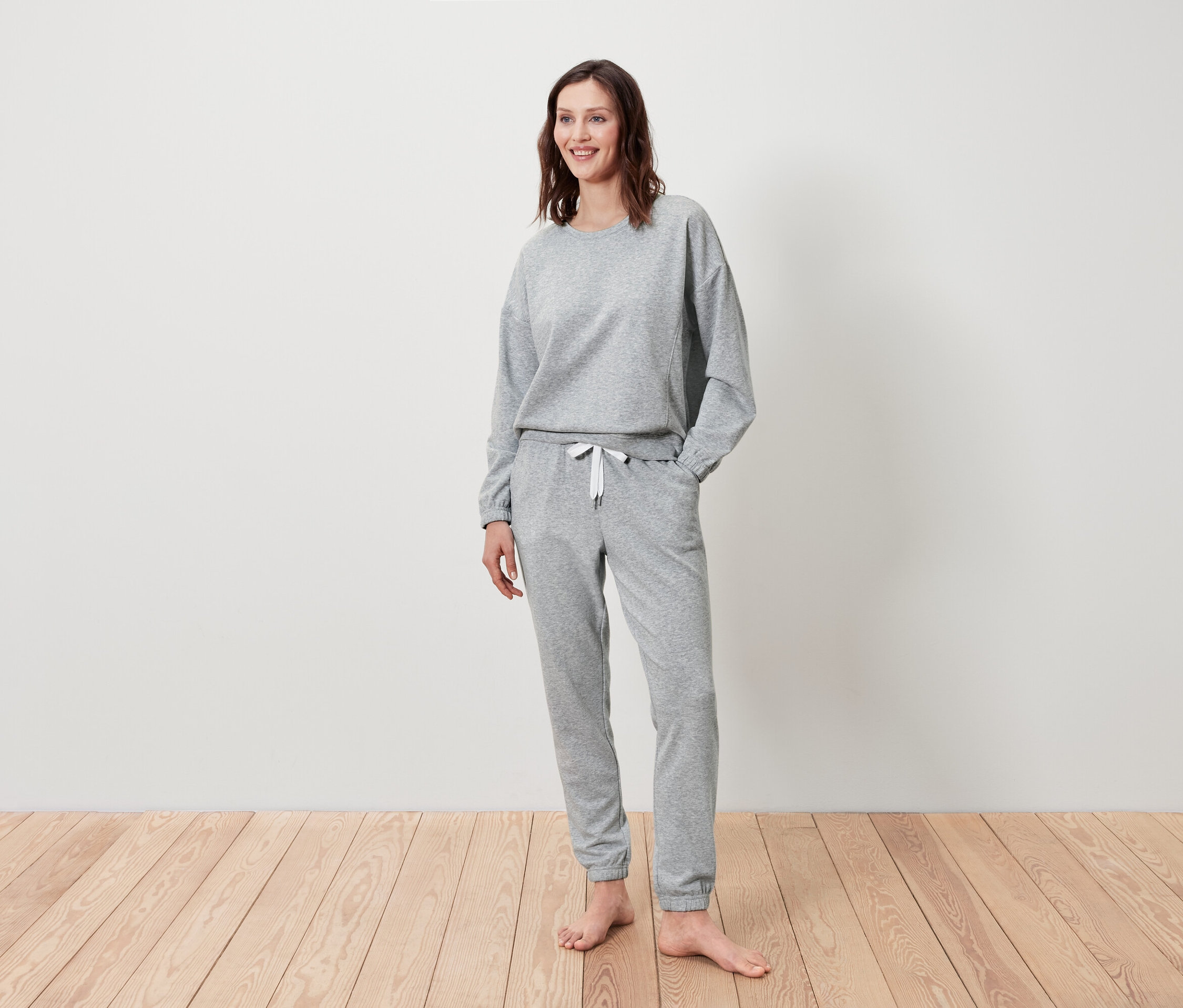 Žena stojí a usmívá se, má na sobě šedou mikinu a tepláky.