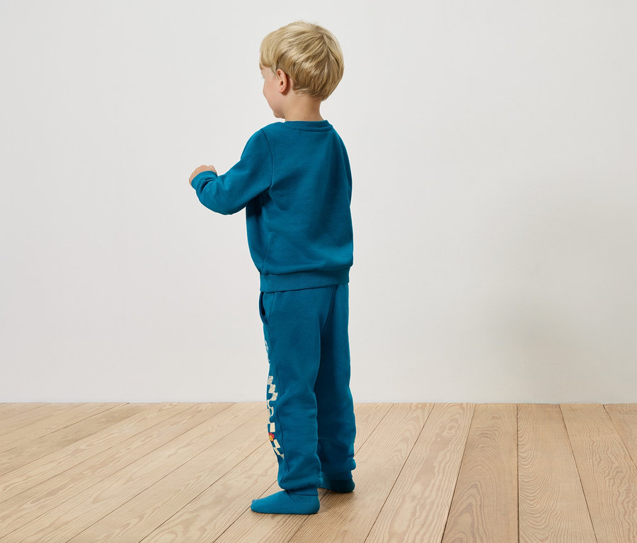 Chlapec v tyrkysové soupravě stojí bokem na dřevěné podlaze.