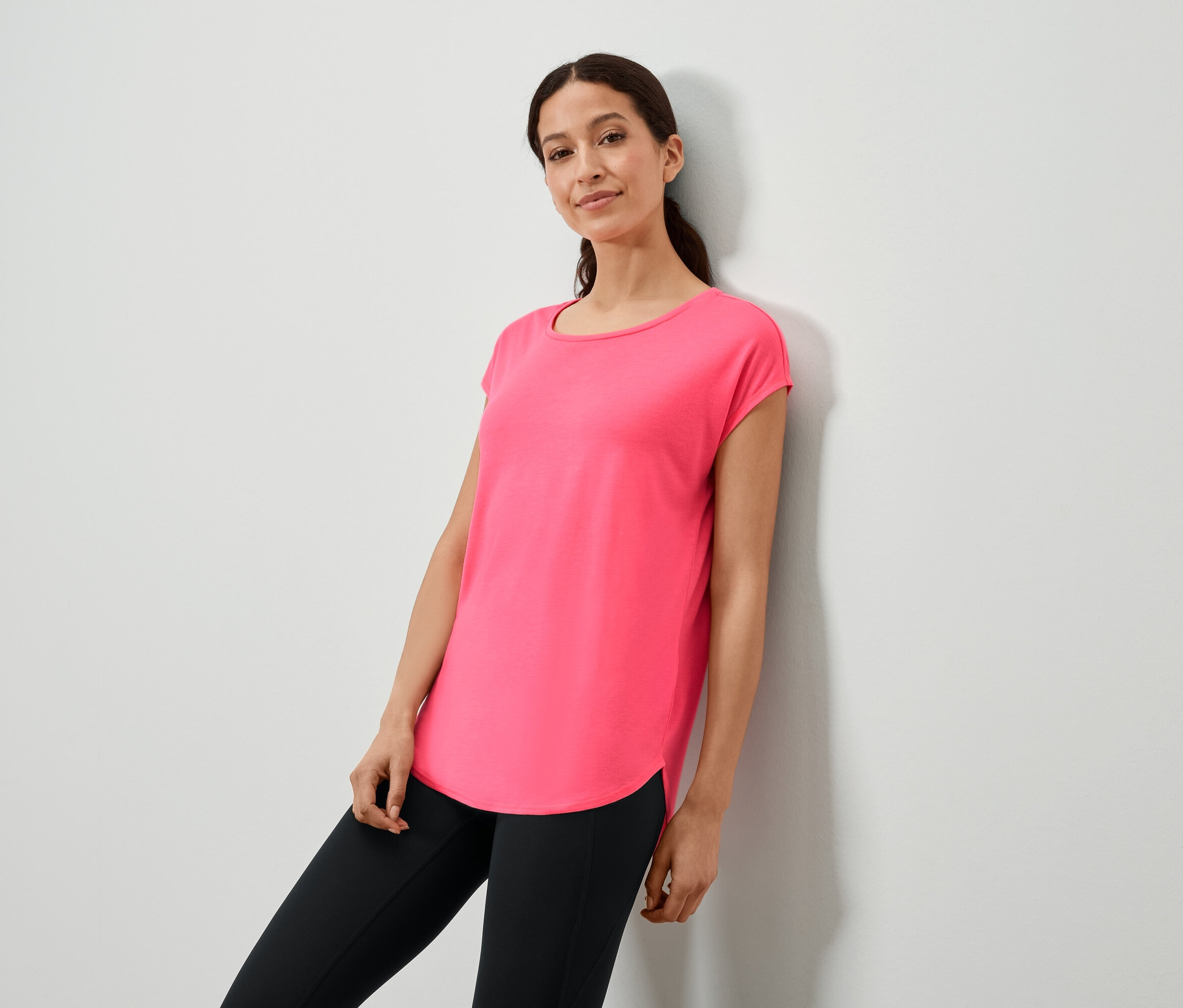 Žena opřená o zeď v růžovém tričku a černých legínách.
