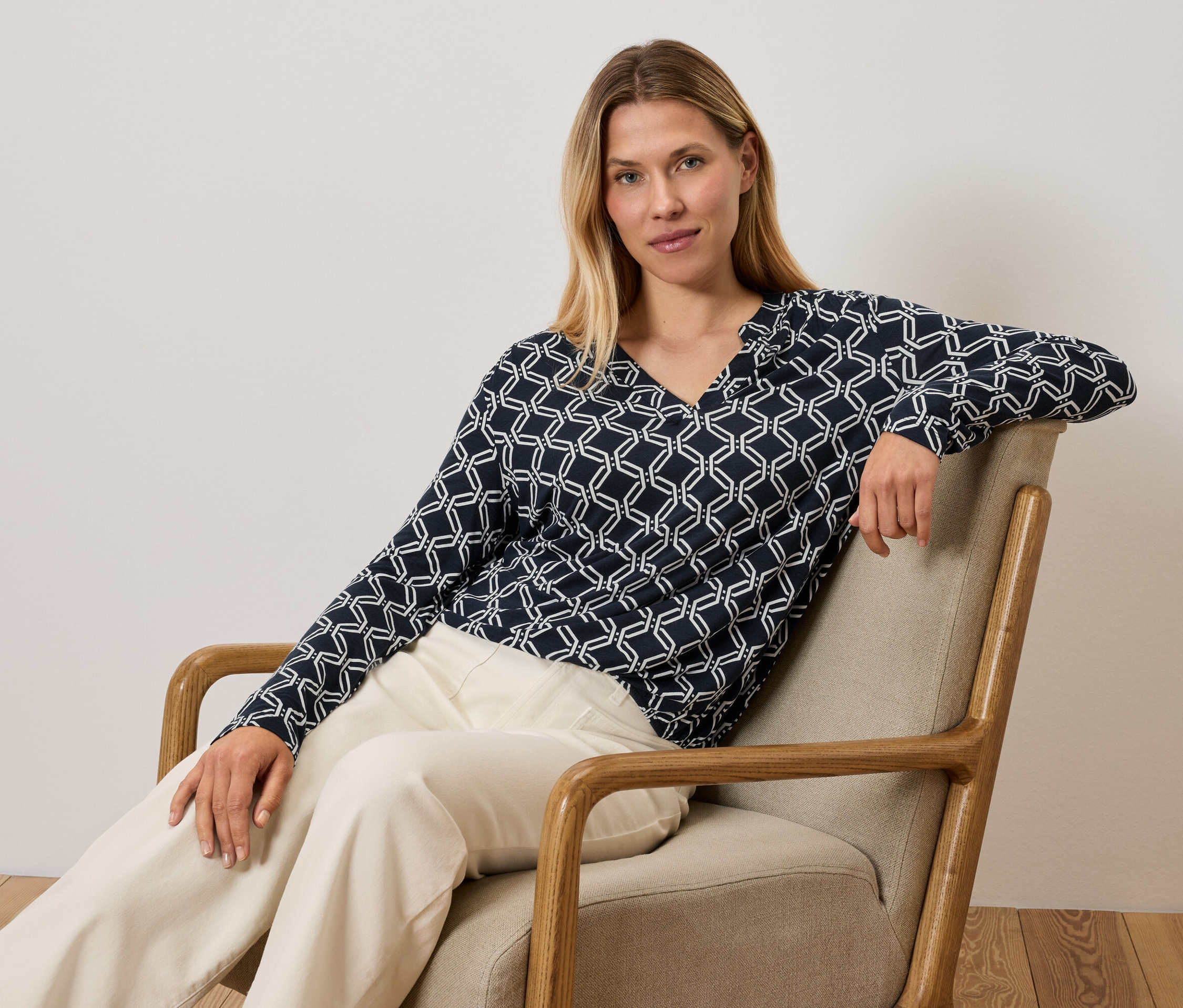 Žena sedí v křesle a má na sobě modro-bílý top a krémové kalhoty.