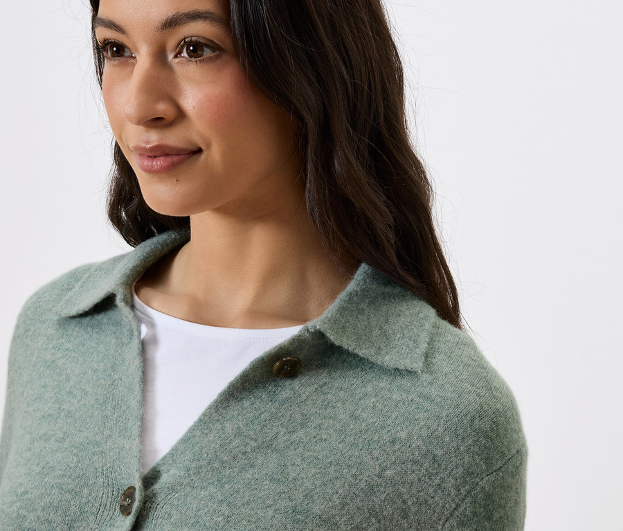 Portrét ženy s tmavými vlasy v zeleném svetru s límečkem a knoflíky.