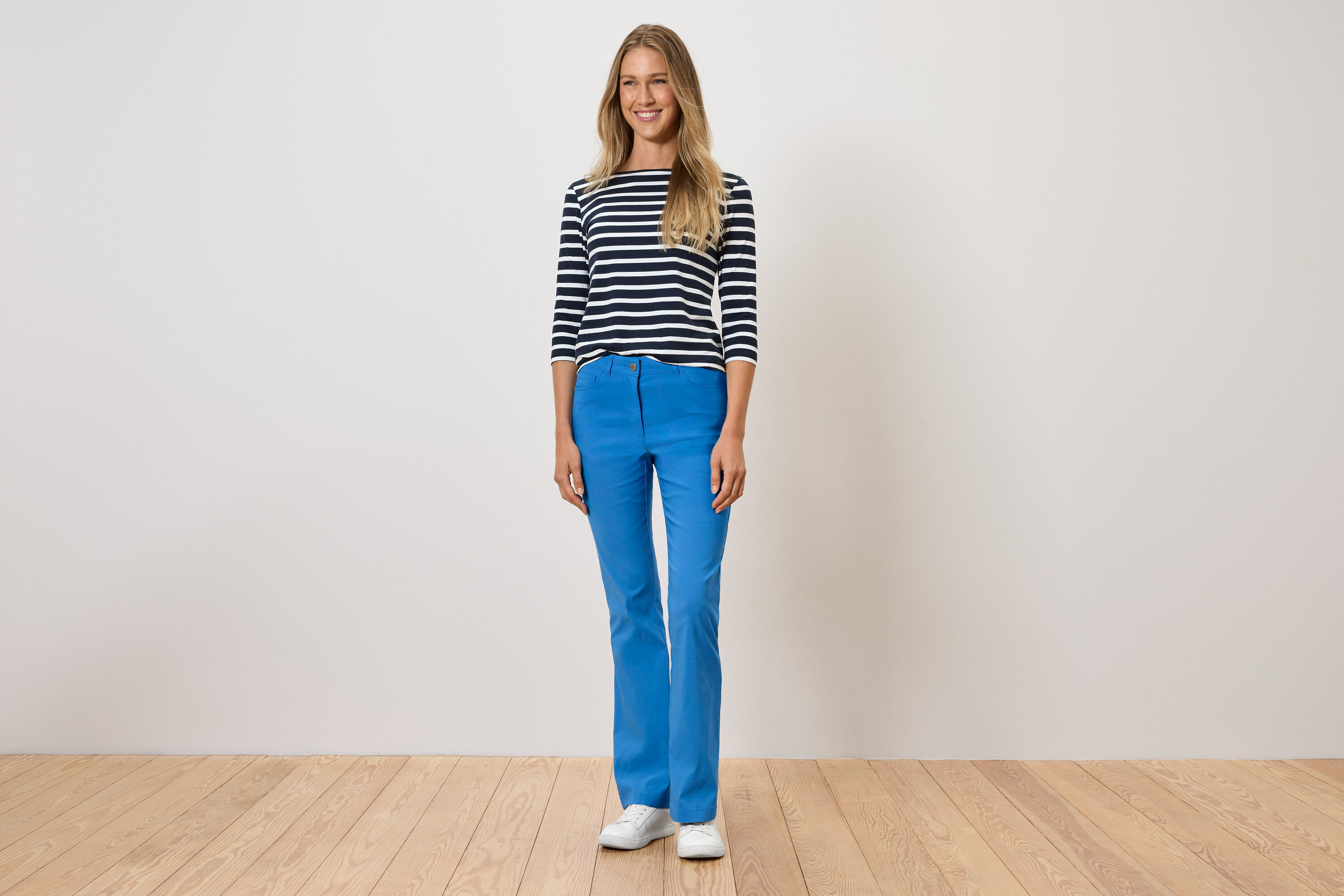 Žena stojí a má na sobě modré kalhoty, pruhované tričko a bílé boty.